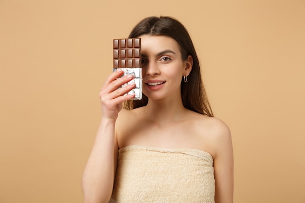 Cerca de una mujer medio desnuda con una piel perfecta, maquillaje desnudo tiene barra de chocolate aislada en la pared de color beige pastel