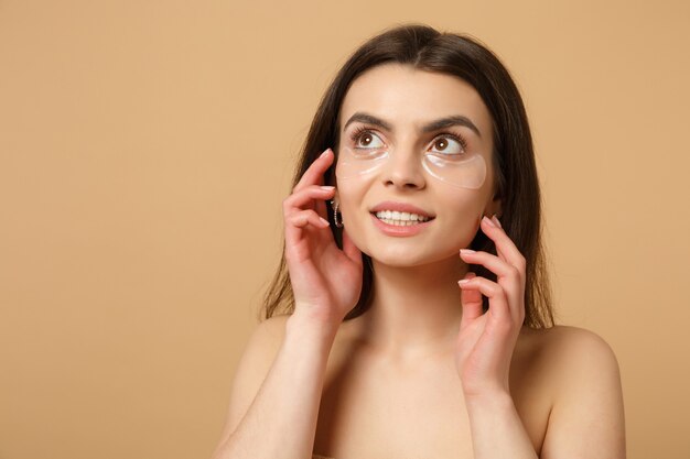 Cerca de una mujer medio desnuda con una piel perfecta, parches de maquillaje desnudos debajo de los ojos aislados en la pared beige pastel