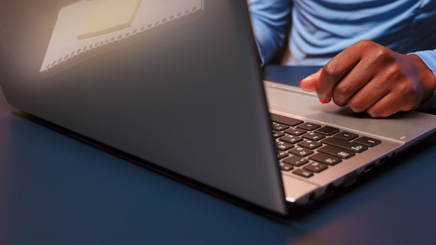 Cerca de mujer negra escribiendo en el teclado del portátil sentado en el escritorio en la sala de estar horas extraordinarias. Empresario africano que trabaja tarde en la noche en el lugar de trabajo personal escribiendo en la computadora.