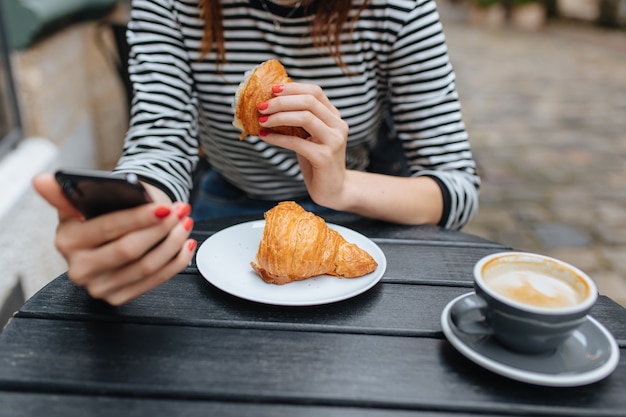 Cerca de mujer en ropa casual con teléfono celular y disfrutando de un café recién hecho con croissant