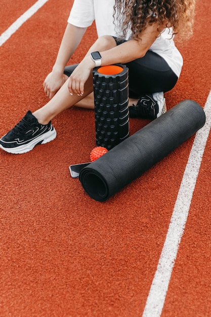 Cerca de una mujer sentada cerca del rodillo mfr, la bola mfr, su teléfono y una alfombra de yoga en una pista de atletismo.