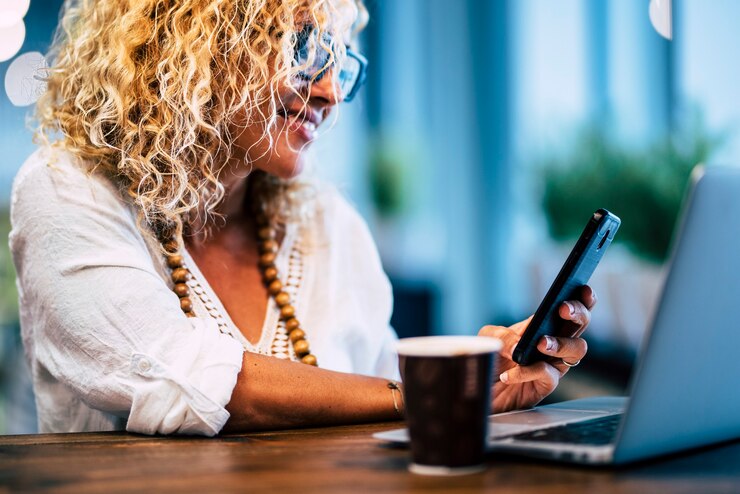 Foto cerca de una mujer sonriendo y mirando su teléfono con su computadora portátil y café