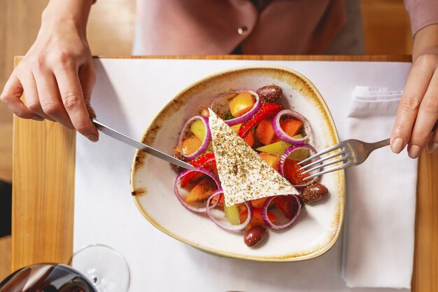 Cerca de mujer sosteniendo tenedor y cuchillo mientras está sentado en la mesa con ensalada fresca con verduras, cebolla y queso