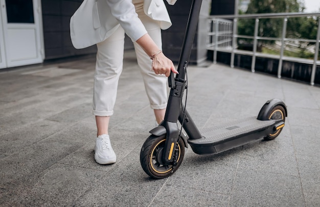 Cerca de una mujer con traje blanco doblando su scooter eléctrico después de un paseo mientras está de pie en el fondo de un edificio moderno