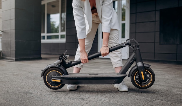 Cerca de una mujer con traje blanco doblando su scooter eléctrico después de un paseo mientras está de pie en el fondo de un edificio moderno