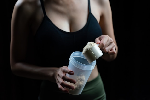 Cerca de mujeres con cucharada de medición de proteína de suero y botella agitadora, preparando batido de proteínas.