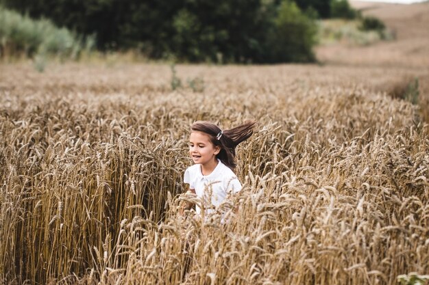 Cerca de niña feliz con largo cabello rubio corriendo a la cámara a través del campo de cebada. Niño sonriente trotar sobre el prado de trigo. Niño lindo pasar tiempo en la plantación de oro. Camara lenta