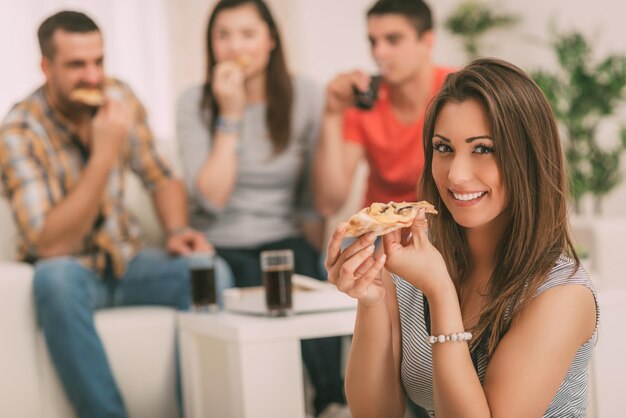 Cerca de una niña sonriendo y comiendo pizza. Sus amigos en el fondo. Enfoque selectivo. Centrarse en la niña, en primer plano.