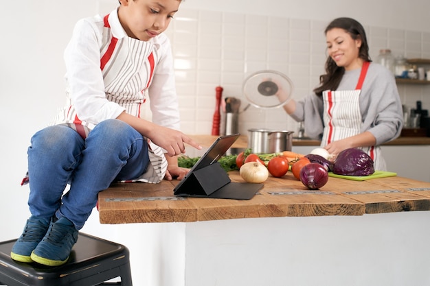 Cerca del niño en el delantal en la cocina jugando con la tableta mientras la madre está cocinando verduras saludables