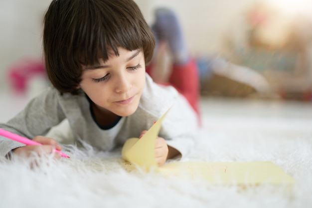 Cerca de niño latino mirando concentrado mientras dibuja con lápices de colores, tirado en el suelo. Niño involucrado en actividad creativa. Aprendizaje a distancia para niños