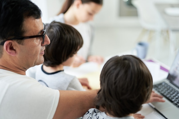 Cerca de padre latino usando laptop mientras trabaja desde casa y mira a los niños. Freelance, encierro, concepto de familia