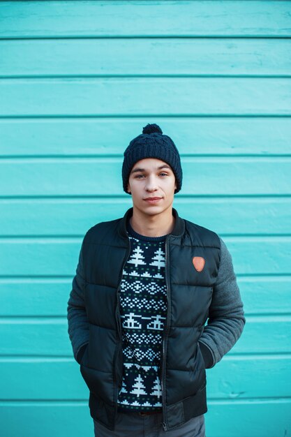 Cerca de la pared azul de madera está parado un joven lindo atractivo con un suéter de punto vintage con un adorno de Navidad con una gorra azul y una elegante chaqueta de invierno. Chico americano encantador.