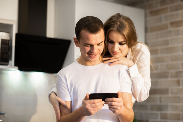 Cerca de una pareja de enamorados usando un teléfono celular juntos en la cocina. Mujer con cabello largo abrazando a un hombre por detrás