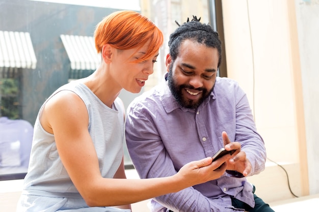 Cerca de la pareja multiétnica sonriendo y mirando al teléfono móvil sentado al aire libre