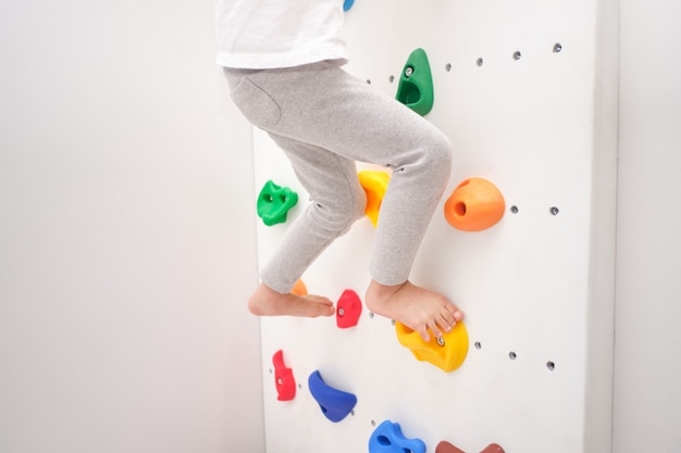 Cerca de las piernas del pequeño niño de jardín de infantes que se divierten tratando de escalar una pequeña pared de roca interior en casa