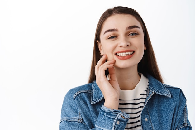 Cerca de retrato de hermosa joven sonriendo con dientes blancos perfectos, tocando la piel limpia y fresca natural, feliz de pie contra la pared blanca en chaqueta de mezclilla para salir