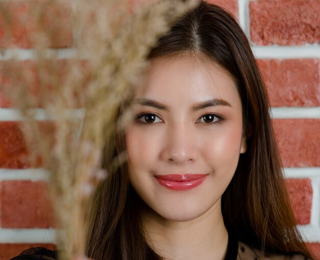 Cerca del rostro de la joven hermosa atractiva modelo femenino asiático sonriendo con rama de flor sobre fondo de pared de ladrillo naranja.