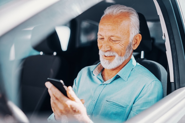 Cerca de senior barbudo sonriente con teléfono inteligente para escribir o leer el mensaje con el brazo en la ventana abierta mientras está sentado en el coche.