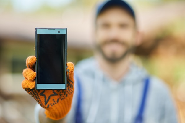 Cerca de smartphone con pantalla en blanco joven constructor masculino en uniforme mediante teléfono móvil