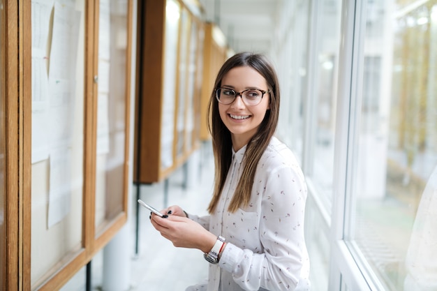 Cerca de sonriente estudiante caucásica con anteojos y cabello castaño con teléfono inteligente