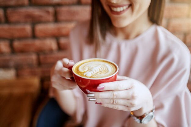 Cerca de sonriente morena con los ojos cerrados, vestida elegante disfrutando de un café en la cafetería