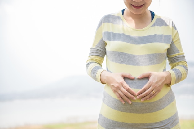 Cerca de la sonrisa de las mujeres embarazadas sosteniendo el estómago, hecho a mano en forma de corazón