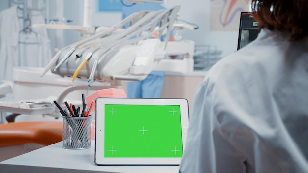 Foto cerca de la tableta digital con pantalla verde horizontal en el escritorio en el gabinete de odontología. dentista analizando plantilla aislada y fondo de maqueta con clave de croma para atención estomatológica