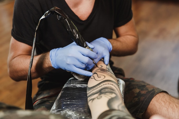 Cerca del tatuador profesional haciendo tatuajes en el brazo de un joven  por máquina con tinta negra