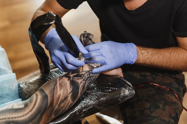 Cerca del tatuador profesional haciendo tatuajes en el brazo de un joven por máquina con tinta negra