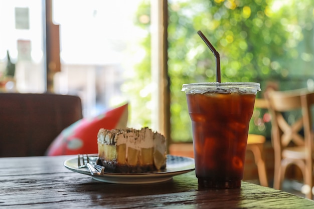 Cerca de una taza de plástico para llevar de café negro helado (Americano) con un trozo de Banoffee sobre la mesa de madera pastel en el restaurante