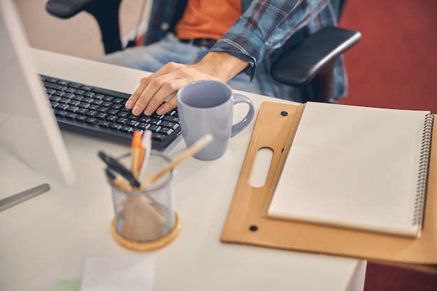 Cerca de trabajador masculino sentado a la mesa con teclado, documentos y taza de café