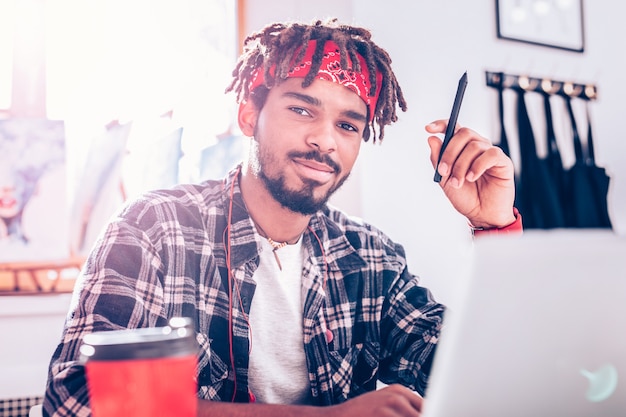 Cerca de la ventana. Artista de pelo oscuro guapo con rastas con diadema roja sentado cerca de la ventana