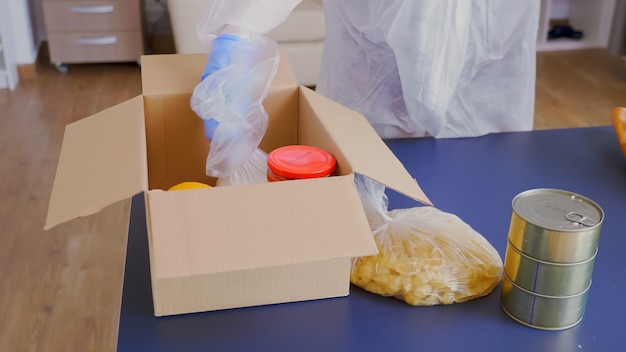 Cerca de voluntarios envasando alimentos en una caja para su entrega vistiendo traje protector durante la pandemia global covid-19
