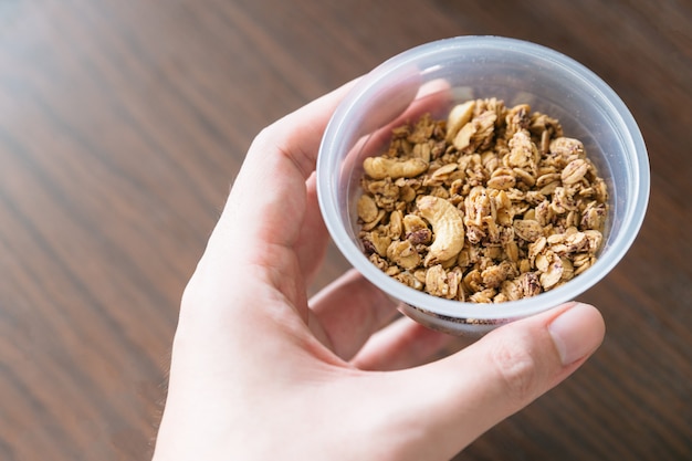 Cereal de granola crujiente orgánico con avena y anacardo dentro de un vaso de plástico con la mano.