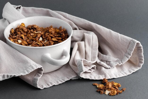 Cereal de granola tostado casero orgánico con chocolate de avena, lino y almendras en un recipiente de vidrio redondo blanco con toalla textil de lino sobre fondo gris Desayuno de muesli natural