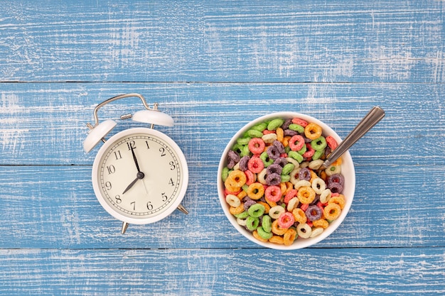Cereales de desayuno de colores en un tazón y despertador blanco sobre un fondo de madera azul concepto de desayuno saludable para niños vista superior