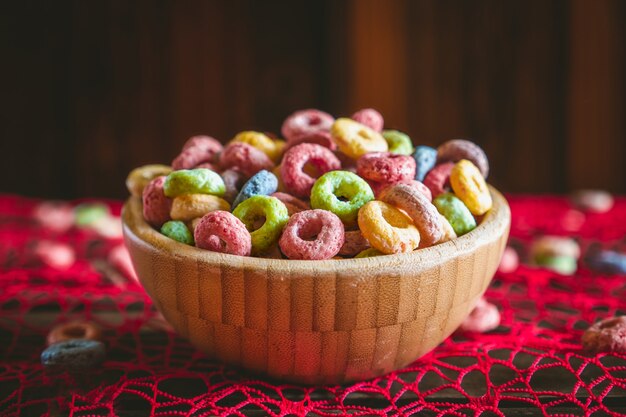 Cereales de frutas redondos coloridos en un cuenco de madera sobre una mesa roja, con algunos esparcidos fuera del cuenco.