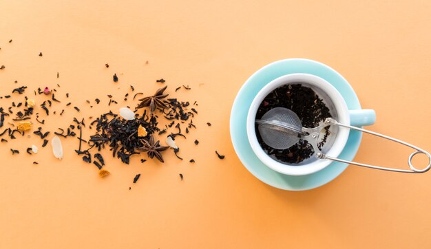 Ceremonia de preparación del té, taza verde menta, colador de té y té seco de hierbas de fruta negra sobre fondo naranja.