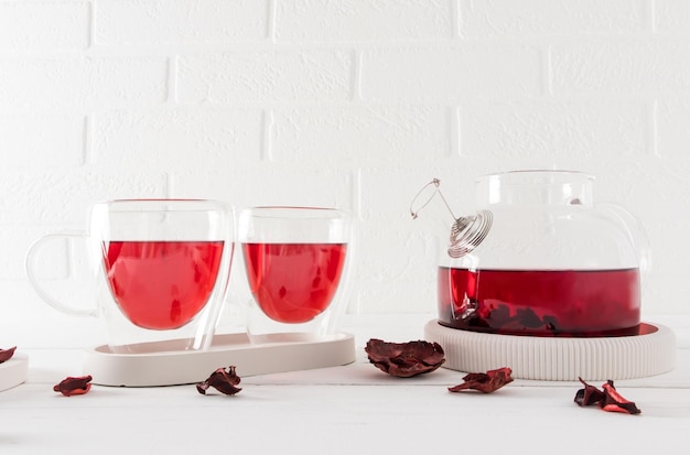 Ceremonia del té una tetera y dos modernas tazas de vidrio de doble fondo con té de rosa de hibisco contra una pared de ladrillo blanco