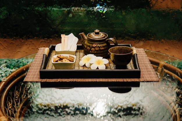 Ceremonia tradicional tailandesa famosa tetera de bronce en bandeja de mimbre con flores de loto, taza, azúcar y galletas en mesa de mimbre con superficie vidriosa con pared de cascada abstracta