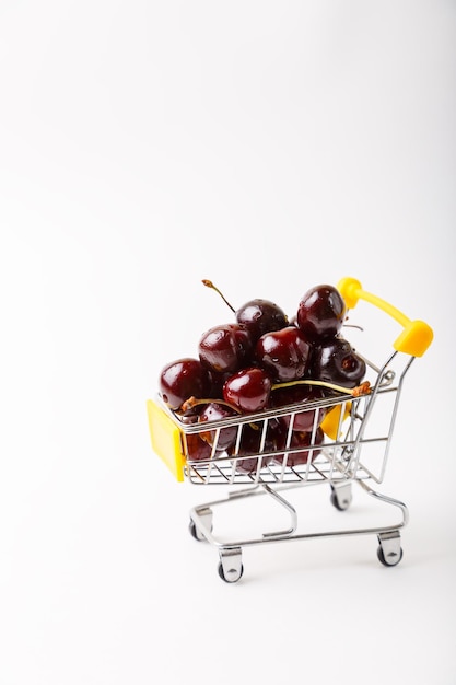 Cereza en un carrito de supermercado pequeño en miniatura para un supermercado Carrito de juguete con bayas sobre un fondo blanco Banner de espacio de copia de primer plano