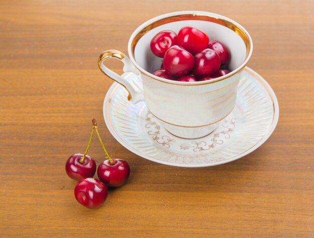 Cerezas dulces en una taza blanca sobre una mesa de madera con dos cerezas en el frente