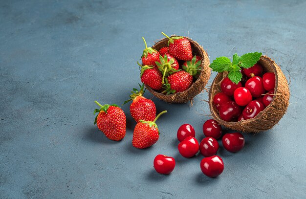 Cerezas y fresas maduras en cáscaras de coco sobre un fondo oscuro. Vista lateral, copie el espacio. Frutas y bayas de verano.