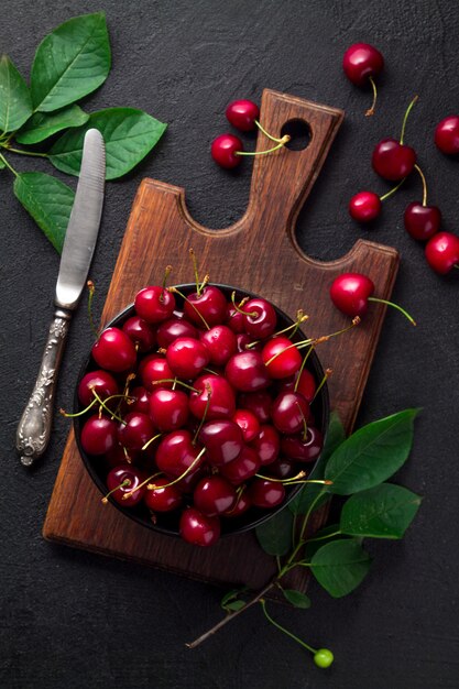 Foto cerezas maduras con hojas verdes en un recipiente negro sobre una tabla de cortar marrón sobre una superficie negra