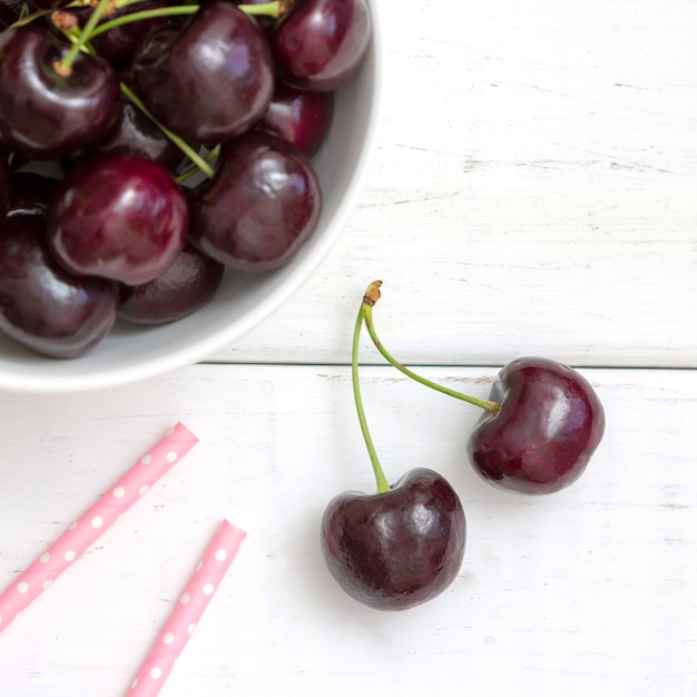 Cerezas negras maduras frescas en un cuenco blanco en una opinión superior del fondo de madera blanco. Lay Flat.