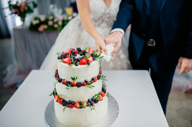 Cerimônia de casamento. Mãos de noivos cortam um bolo de três camadas branco com morangos e amoras