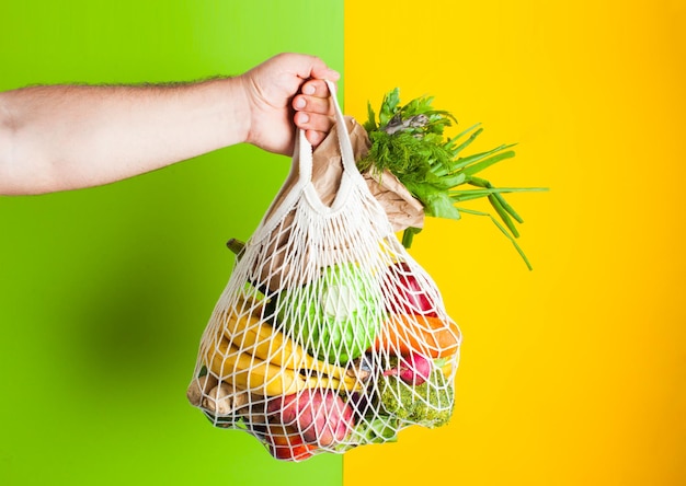 Foto cero desperdicio de compras. la mano del hombre sostiene una bolsa de algodón llena de verduras sobre fondo verde y amarillo. producto en bolsa de papel. lugar para texto, comida vegana.