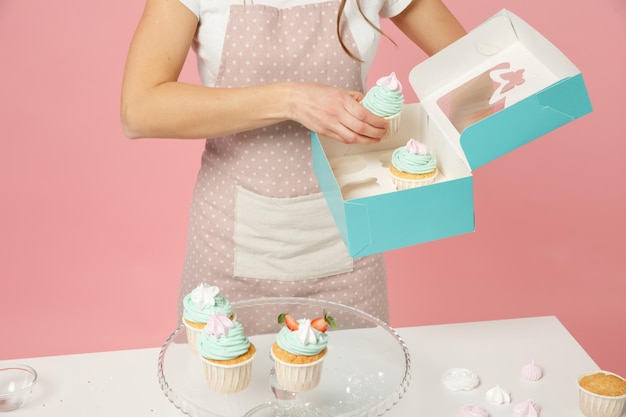 Cerrar ama de casa recortada cocinera repostera o panadera en delantal camiseta blanca embalaje pastel cupcake en mesa aislada sobre fondo rosa pastel en estudio. Simule el concepto de comida en el espacio de copia.