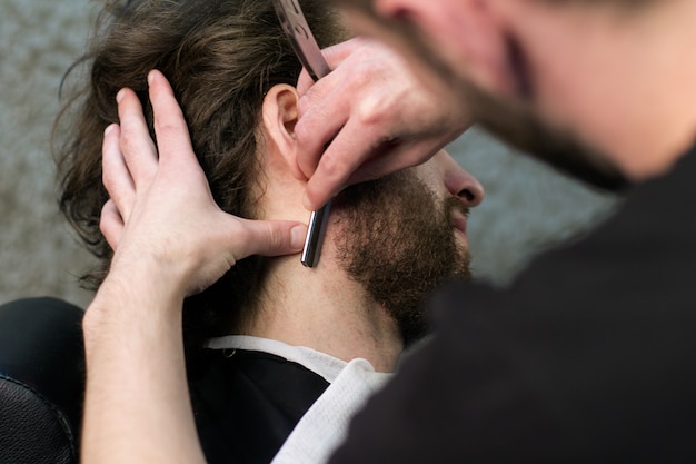Cerrar la cara del hombre en la barbería: las manos del barbero se afeitan la barba con una navaja de afeitar