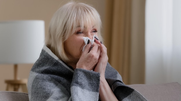 Cerrar enferma vieja abuela rubia 60s mujer cubierta con una manta caliente en casa sonarse la nariz